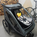 Gray stroller with a child in a car seat and a yellow duck toy, against a background with firewood and a bicycle.