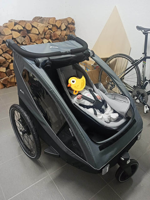 Gray stroller with a child in a car seat and a yellow duck toy, against a background with firewood and a bicycle.